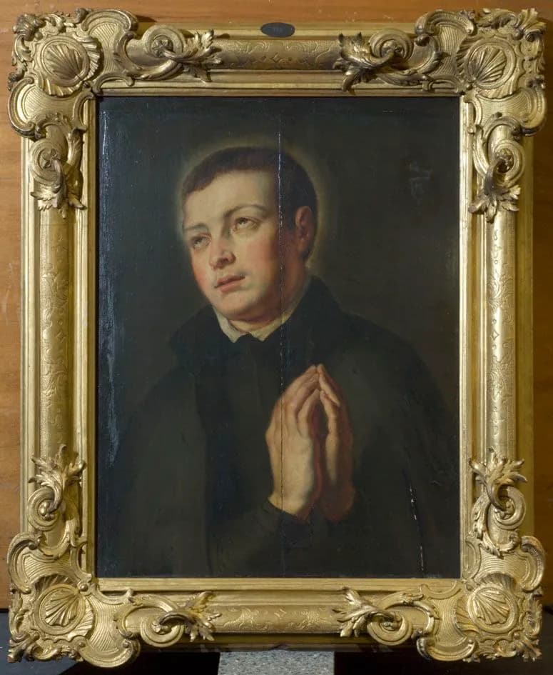 A painting of a young man in a dark Jesuit habit with a white collar, hands joined in prayer and eyes raised, a faint halo behind his head, in an ornate gilt frame