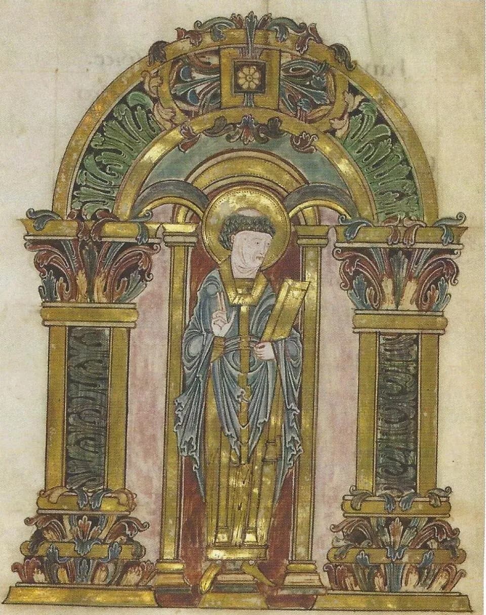 A manuscript painting of a tonsured bishop with a gold halo, wearing blue vestments, raising his right hand in blessing and holding a closed book, standing beneath a gilded arch between two ornamented columns