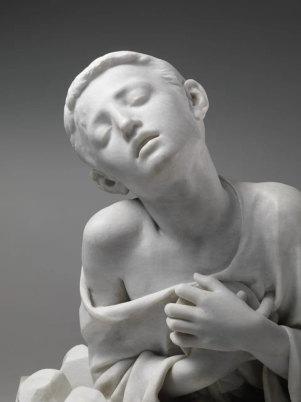 Detail of a white marble sculpture of a dying boy with short hair, his eyes closed and his head fallen to one side, both hands pressing a hidden object against his chest beneath his tunic, with stones lying beside him