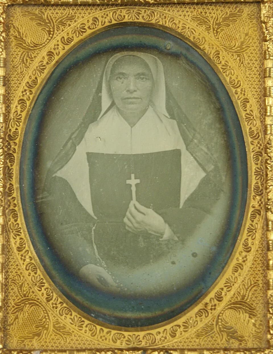 A daguerreotype of Mother Théodore Guérin in an oval gilt frame, an older woman in a white bonnet and collar under a dark veil, holding a crucifix against her chest