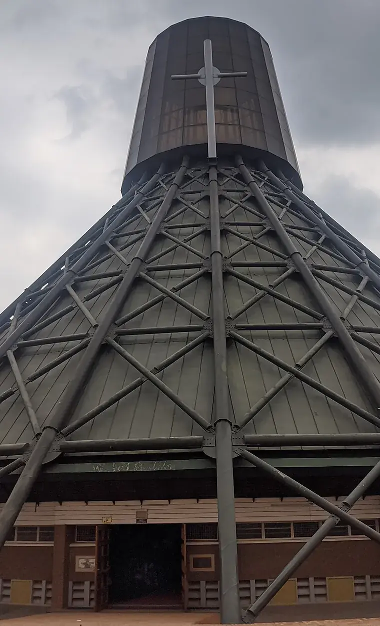 The Basilica of the Uganda Martyrs at Namugongo, seen from the front under a gray sky: a tall cone of green metal roofing braced by a lattice of steel tubes, rising to a dark cylindrical tower with a large metal cross, above a low brick wall with an open doorway