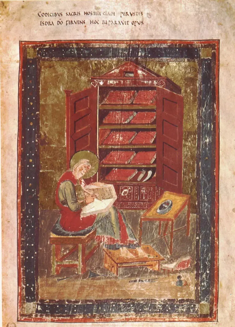 Early medieval manuscript painting of an old bearded scribe with a gold halo, seated on a stool writing in a book on his knees, before a red cupboard with open doors and shelves of bound books, inside a decorated border, with two lines of Latin above naming Esdra
