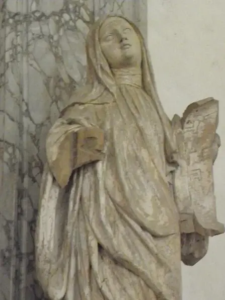 Carved and whitened wooden statue of a veiled nun in a full habit and mantle, looking upward and holding a book in her left hand, in the collegiate church at Champeaux
