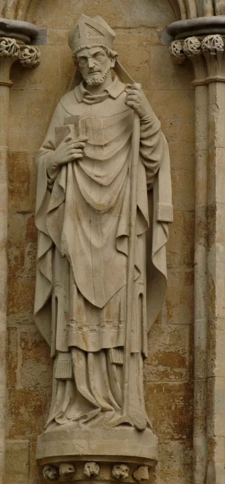 Weathered stone statue of a bearded bishop in a mitre and vestments, standing in a niche with two books held to his chest in one hand and a staff in the other, on the West Front of Salisbury Cathedral